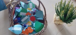 Koszyk wiklinowy z pamiątkowymi medalami wydrukowanymi w 3D. Obok koszyka stoi doniczka z zieloną rośliną.