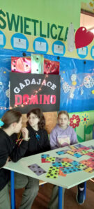 Trzy uczennice siedzące przy stoliku na którym widać kartki z symbolami gry w domino. W tle widać , plakat z napisem gadające domino