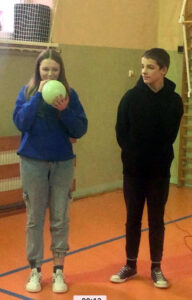 Uczeń i uczennica stoją obok siebie. Uczennica próbuje nadmuchać zielony balonik.