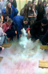 Prowadzący eksperyment, wylewa substancję która na podłodze zamienia się w dym. Wokół prowadzącego stoi grupa uczniów.