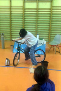 Uczennica bierze udział w pokazie na sali gimnastycznej. Próbuje jechać na rowerze o nietypowej konstrukcji.