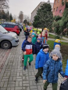 Grupa przedszkolaków spaceruje wzdłuż chodnika. Na zdjęciu widać dzieci niosące kukłę Marzanny