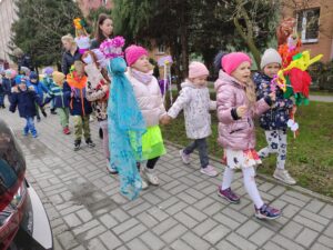 Grupa przedszkolaków spaceruje wzdłuż chodnika. Na zdjęciu widać dzieci niosące kukłę Marzanny